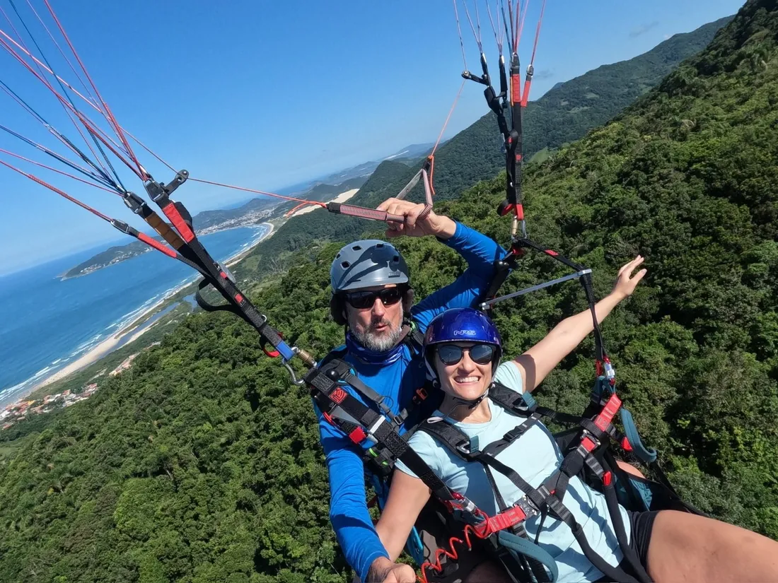 Voo duplo de parapente sobre o litoral, com a praia e o morro verde lá embaixo